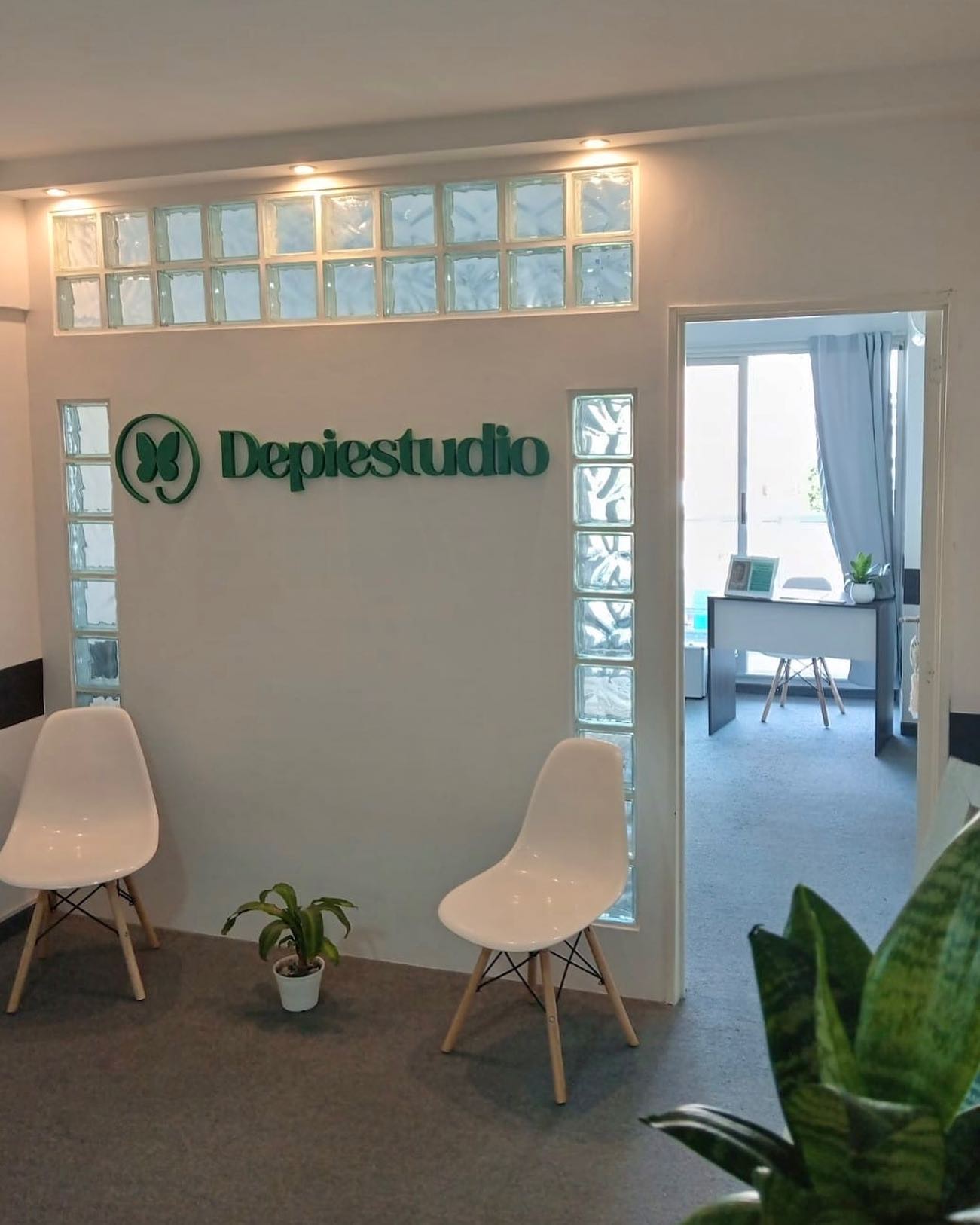 La recepción de Depiestudio: el cartel verde con el logo de la mariposa, ladrillos de vidrio, sillas blancas, plantas y el consultorio al fondo